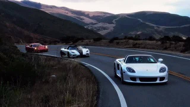 Porsche Carrera GT, Koenigsegg and Ferrari following a winding road through coastal hills